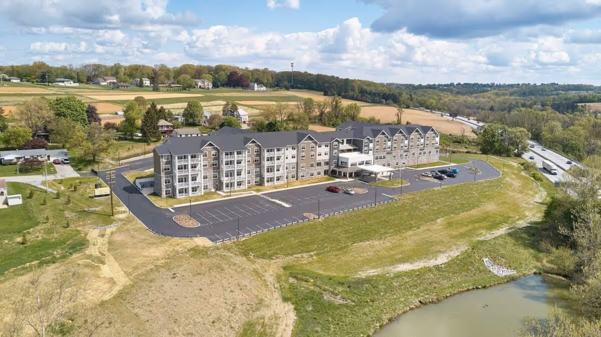 Aerial view of a large, multi-story senior living facility named The Residence at Fitz Farm, surrounded by open fields, trees, and a small pond. The building has a modern design with multiple balconies and a spacious parking lot with several cars parked. A highway is visible in the background under a partly cloudy sky.