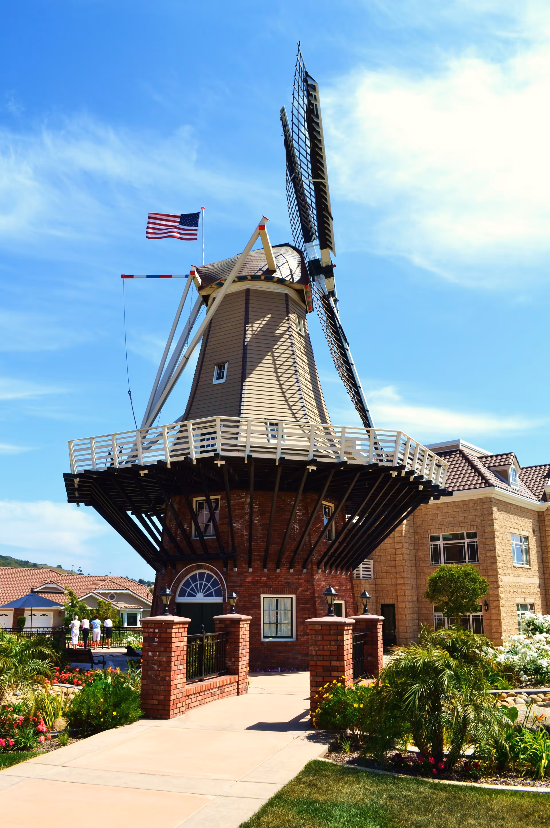 A decorative windmill tower with large blades and an American flag in front of a landscaped building under a blue sky.