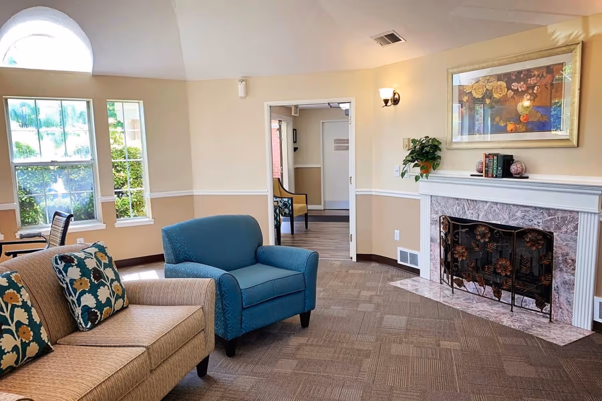 Bright communal living room with upholstered chairs, a fireplace, artwork, and windows.