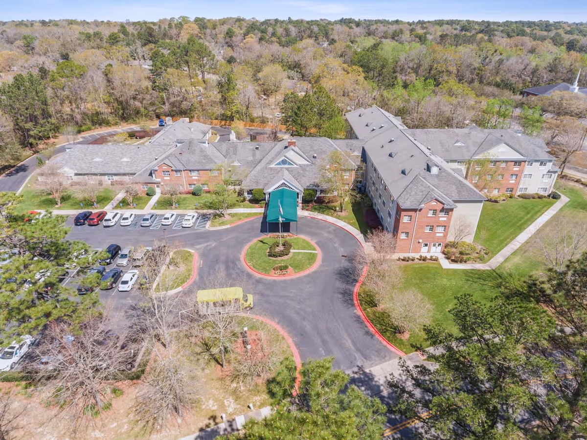 Aerial view of a multi-wing senior living building with a covered entrance, circular driveway, parked cars, and surrounding trees.