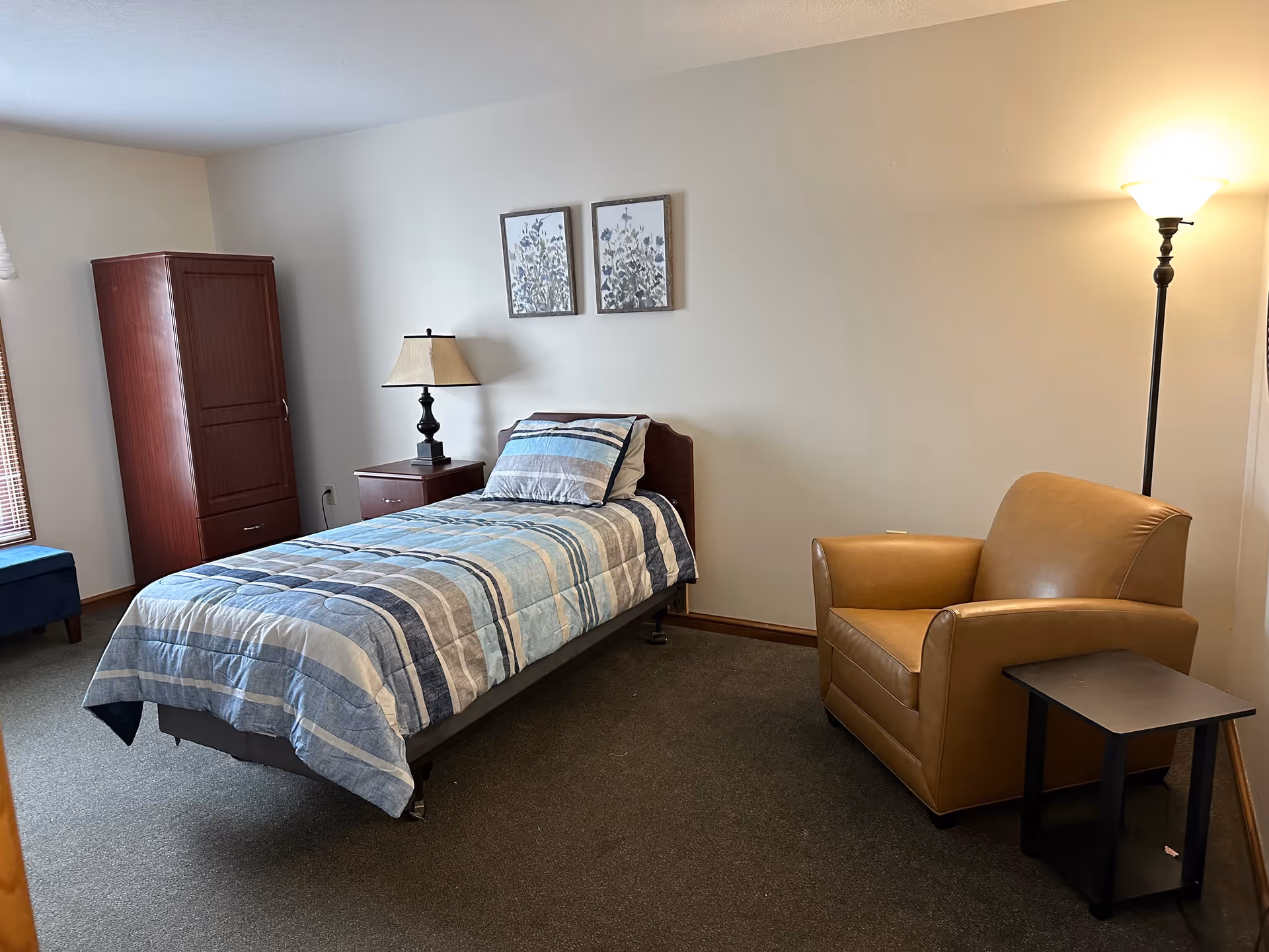 A neatly arranged bedroom with a single bed covered in a blue and gray striped comforter. Next to the bed is a wooden nightstand with a lamp. On the right side of the room is a tan leather armchair with a small black side table beside it. There is a tall wooden wardrobe against the wall near a window with blinds. Two framed botanical prints hang on the wall above the bed. The room has beige walls and a carpeted floor.