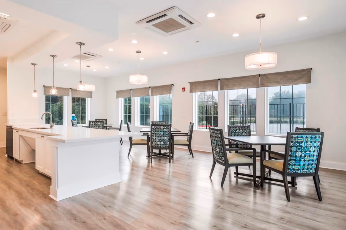 Bright communal dining area with round tables and patterned chairs adjacent to a white kitchen island and large windows.