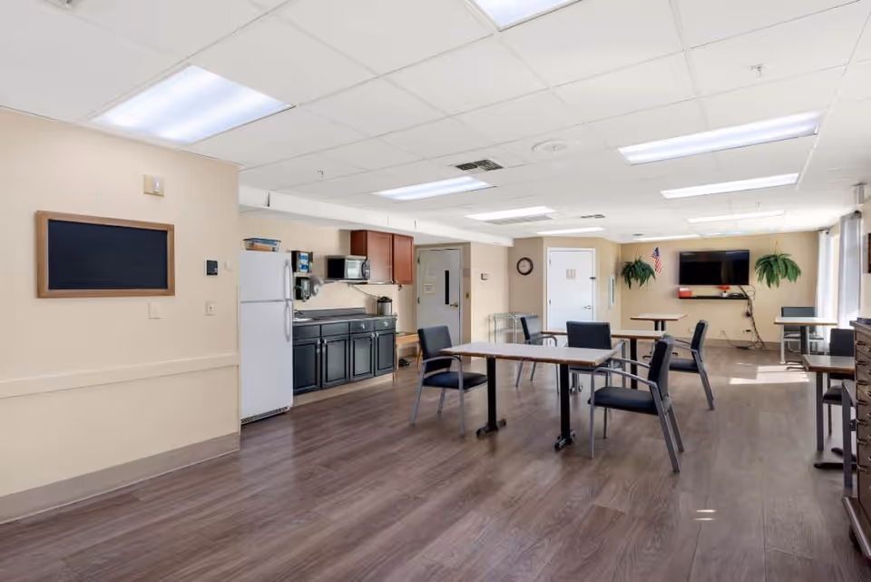 A spacious common area with several tables and chairs arranged for seating. The room has a kitchenette with a refrigerator, microwave, and cabinets along one wall. There is a wall-mounted TV, two hanging plants, and a clock on the walls. The floor is wood, and the ceiling has fluorescent lighting panels.