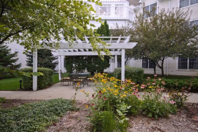 Outdoor garden area with a white pergola over a concrete patio. There are metal chairs and a table under the pergola. Surrounding the patio are green bushes, trees, and colorful flowers. The background shows the exterior of a white building with multiple windows.