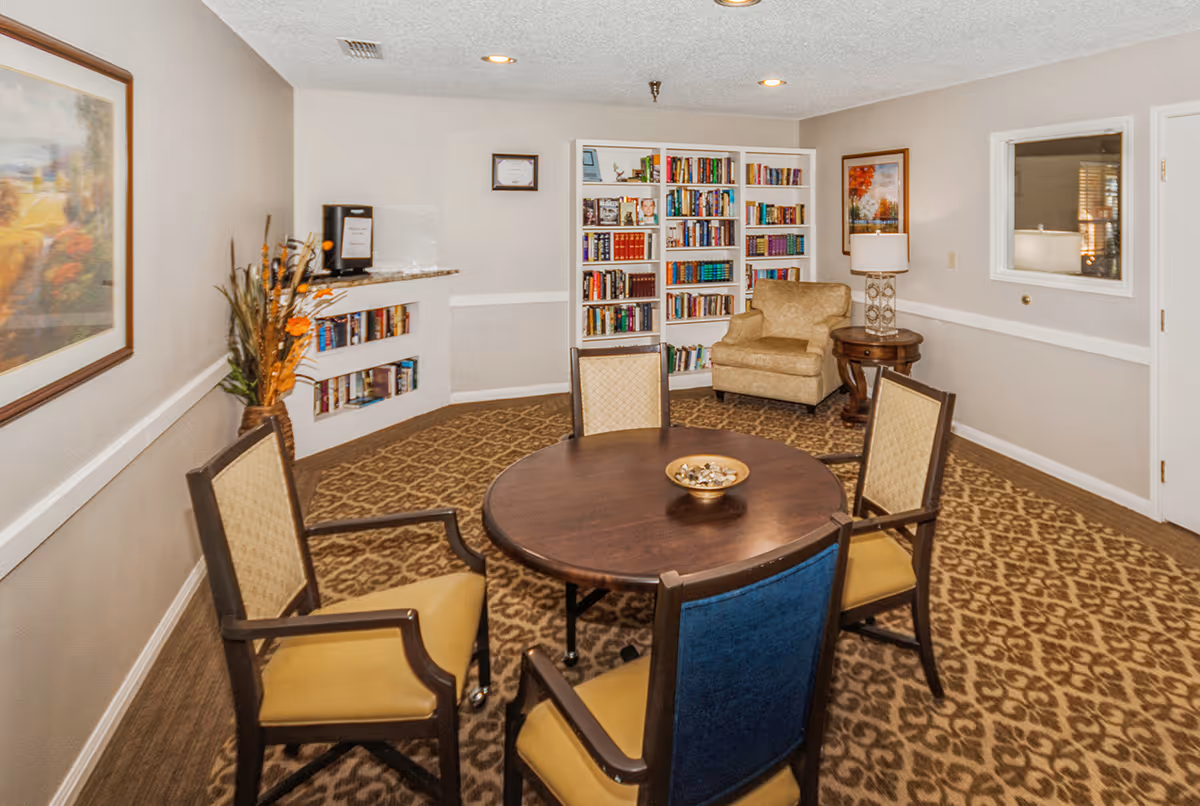 A cozy interior room with a round wooden table surrounded by four chairs, a beige armchair next to a small round wooden side table with a lamp, a white bookshelf filled with books, and framed artwork on the walls. The room has patterned carpet and neutral-colored walls.