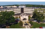 Front exterior of a curved multi-story senior living building with a covered entrance, landscaped courtyard and parked cars.