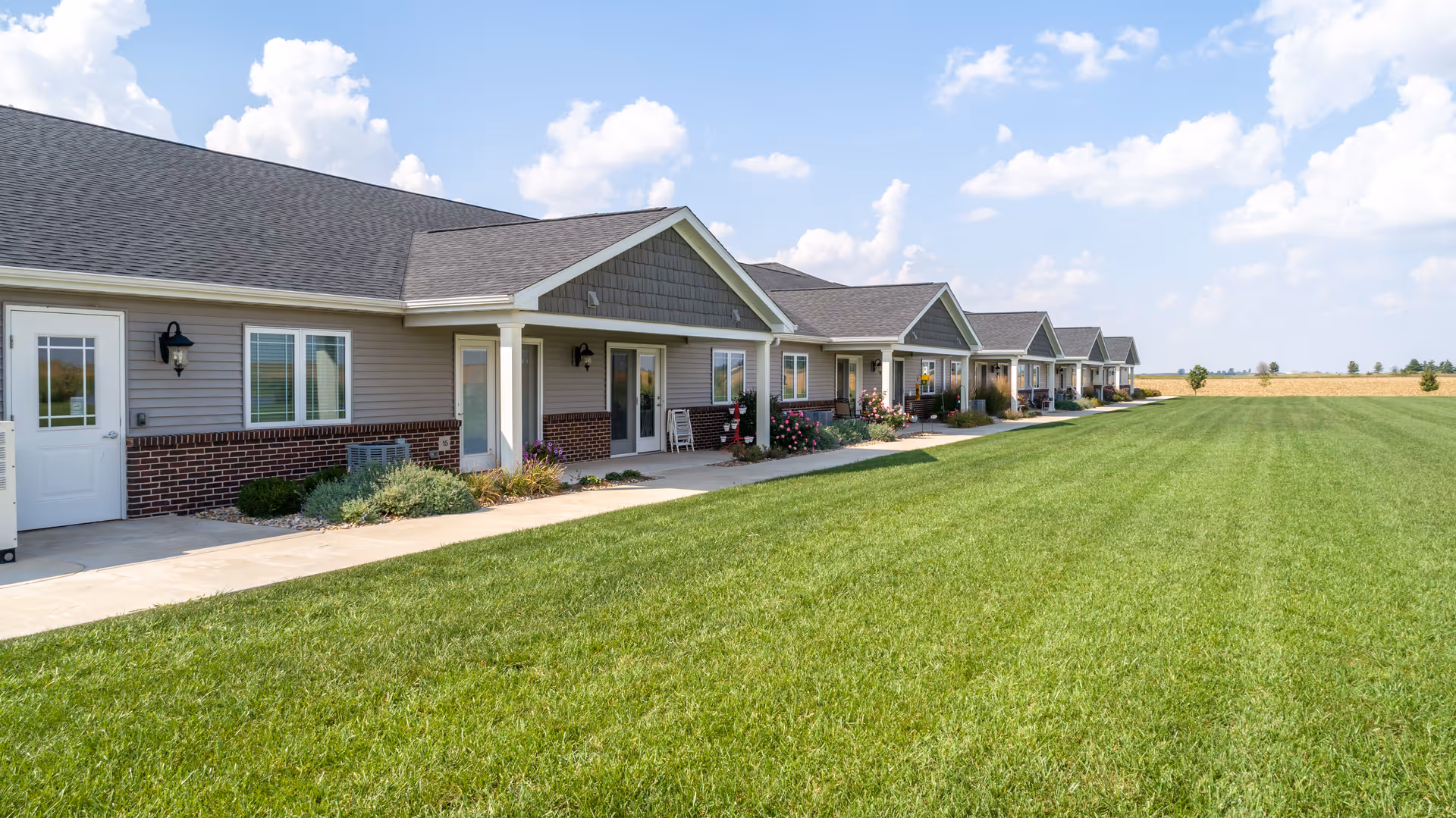 Exterior view of a single-story senior living facility building with multiple units, each having a small porch area. The building features gray siding with brick accents and a dark shingled roof. A large, well-maintained green lawn extends in front of the building under a partly cloudy sky.