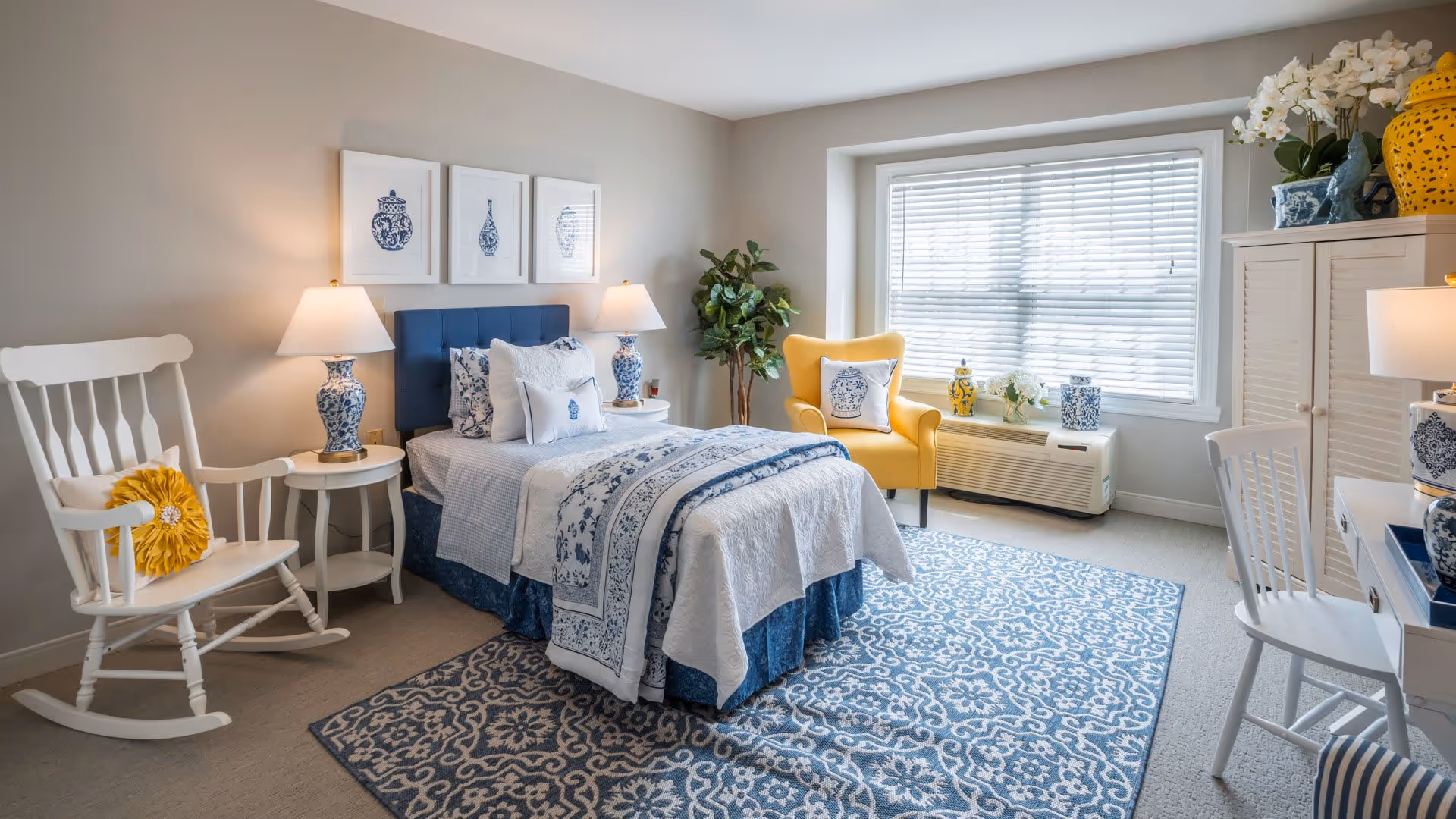 A bright and cozy bedroom with a single bed dressed in blue and white bedding, flanked by two matching blue and white lamps on white nightstands. There is a white rocking chair with a yellow cushion on the left, a yellow armchair with a decorative pillow near the window, and a white desk with a chair on the right. The room features a large patterned blue and white rug, three framed blue and white prints above the bed, and various blue and white decorative items throughout.