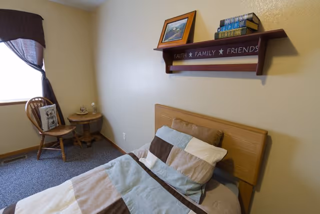 Small bedroom with a single bed and striped bedding, wooden headboard, a wall shelf reading 'Faith Family Friends', and a chair and side table by a curtained window.