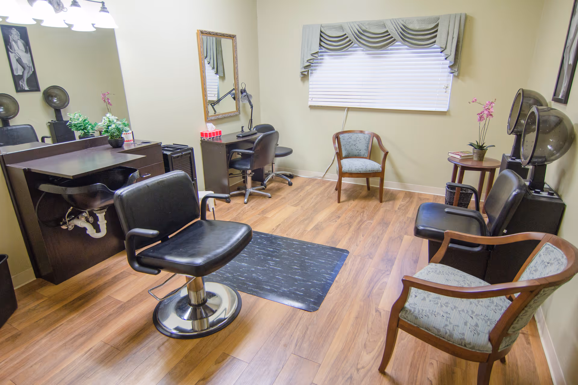 Interior of a hair salon room with wooden flooring, featuring a black salon chair in front of a sink and counter with plants, a mirror on the wall, two hair drying stations with black chairs, and additional seating with upholstered chairs. A window with blinds and a valance is visible on the far wall.