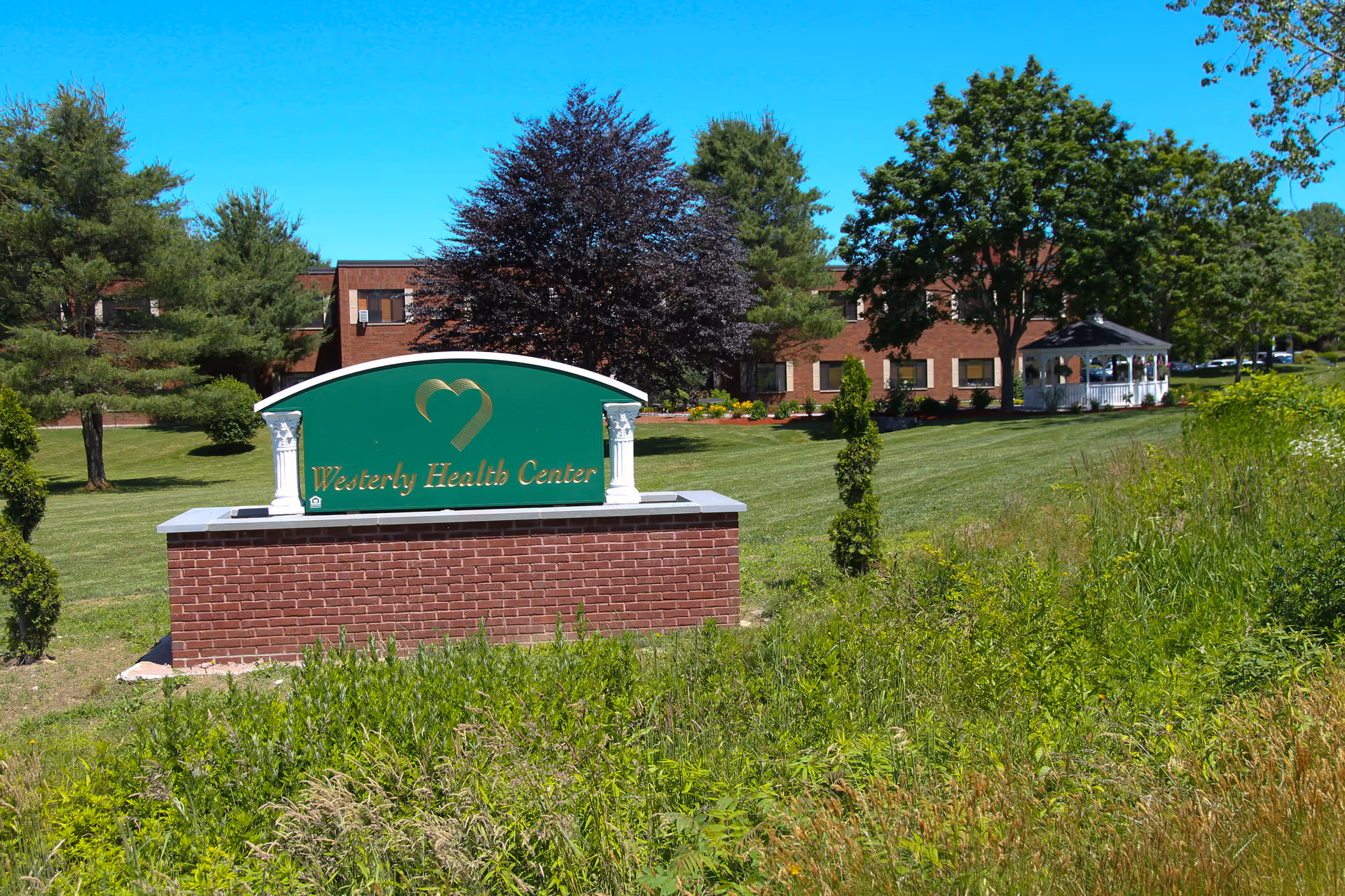 Outdoor view of Westerly Health Center with a green sign featuring a gold heart logo and the facility name. The background shows a large brick building, green lawn, trees, and a white gazebo under a clear blue sky.