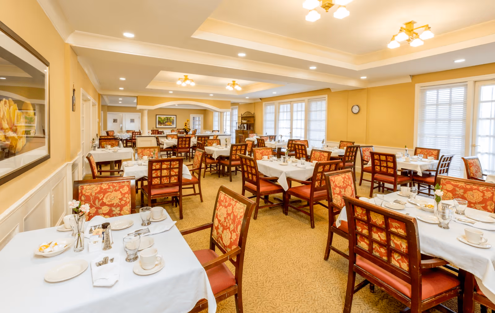A bright and spacious dining room with multiple tables covered in white tablecloths, each set with plates, cups, glasses, and silverware. The chairs have wooden frames with red and gold patterned cushions. The room has beige walls, large windows with blinds, and ceiling lights providing warm illumination. There are framed pictures on the walls and a clock on one side.
