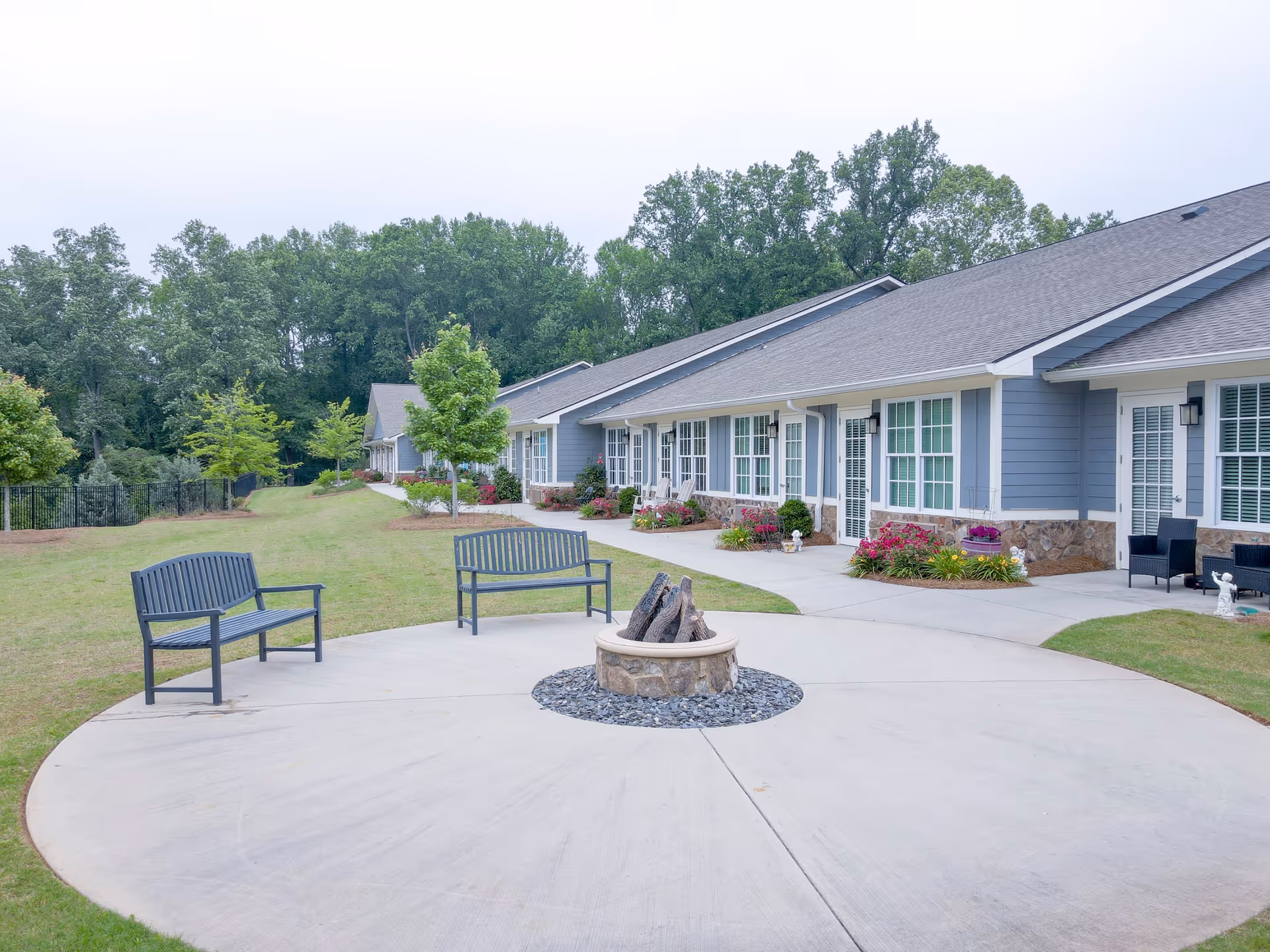 Outdoor patio area at Manor Lake Assisted Living & Memory Care - Gainesville featuring a circular concrete seating area with a fire pit in the center, surrounded by two metal benches. The building exterior is visible with blue siding, white trim, and multiple windows and doors. There are landscaped flower beds and trees along the walkway, with a grassy lawn and wooded area in the background.