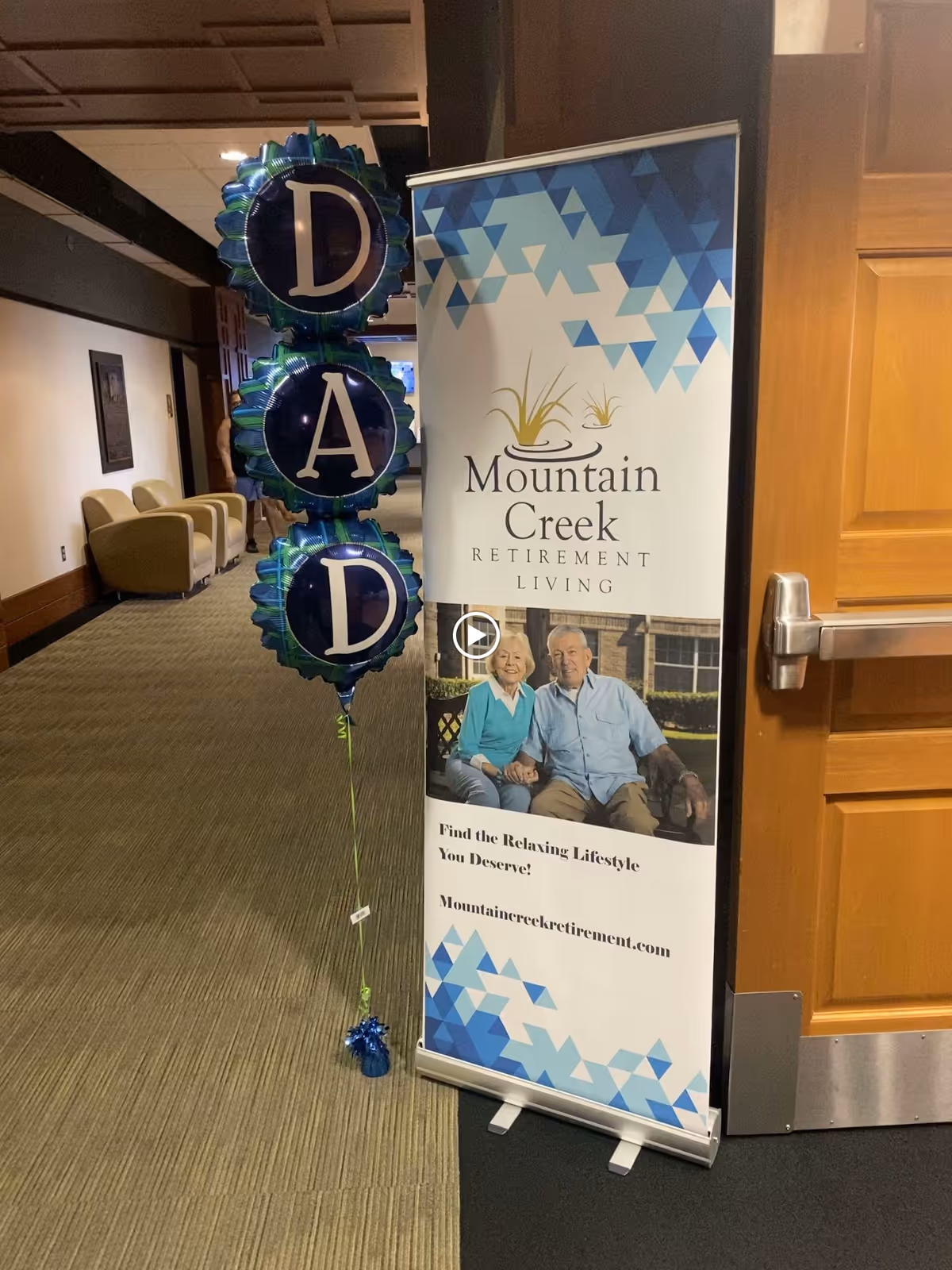 Indoor hallway of a retirement living facility with a standing banner for Mountain Creek Retirement Living featuring an elderly couple sitting and smiling. Next to the banner are three blue balloons stacked vertically spelling out 'DAD'. There are chairs along the hallway wall and a wooden door partially visible on the right.
