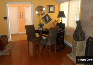 A cozy interior room with wooden flooring, featuring a small table with two wicker chairs. The wall is decorated with three mirrors of different shapes. There is a lamp on a side table near a window with sheer curtains. A large decorative vase stands next to a stone fireplace.