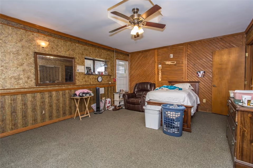 Spacious bedroom with a bed, recliner, dresser, laundry basket, and wood-paneled walls.