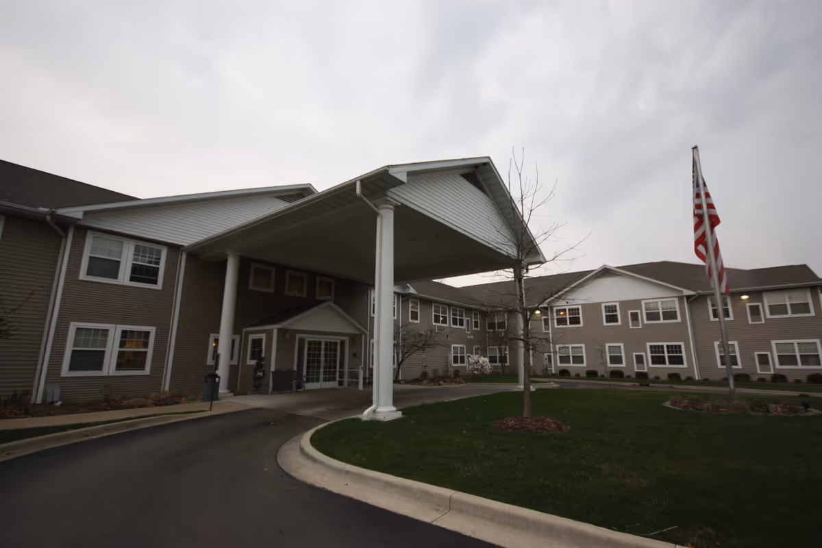 Exterior view of Eagle Ridge of Decatur senior living facility showing a two-story building with beige siding, white trim, and multiple windows. The entrance features a covered driveway supported by white columns. There is a small landscaped area with a tree and an American flag on a flagpole near the entrance.