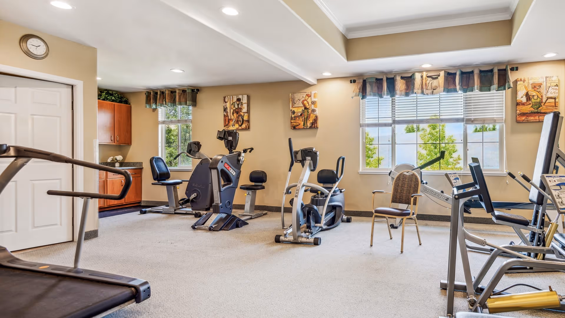 A well-lit exercise room with various fitness equipment including stationary bikes, a treadmill, and a rowing machine. The room has beige walls, large windows with blinds and valances, and framed artwork on the walls. There is a clock above a white door and a wooden cabinet with a countertop in the corner.