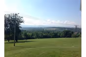 A wide grassy lawn sloping toward distant tree-covered hills and a clear blue sky with utility poles in view.