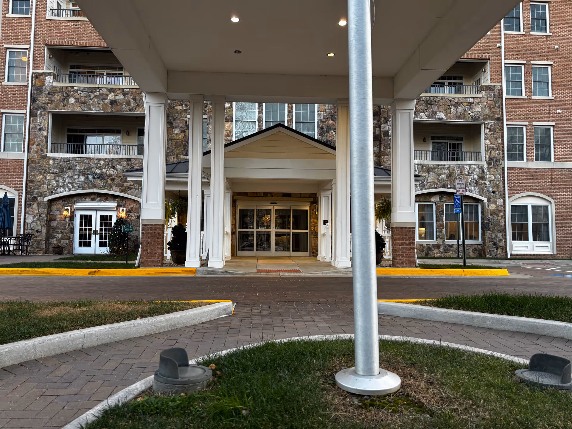 Entrance of a multi-story senior living facility with a covered drop-off area supported by white columns. The building exterior features a combination of red brick and stone accents, with multiple windows and balconies. There is a paved driveway and a small grassy area with a flagpole in front.