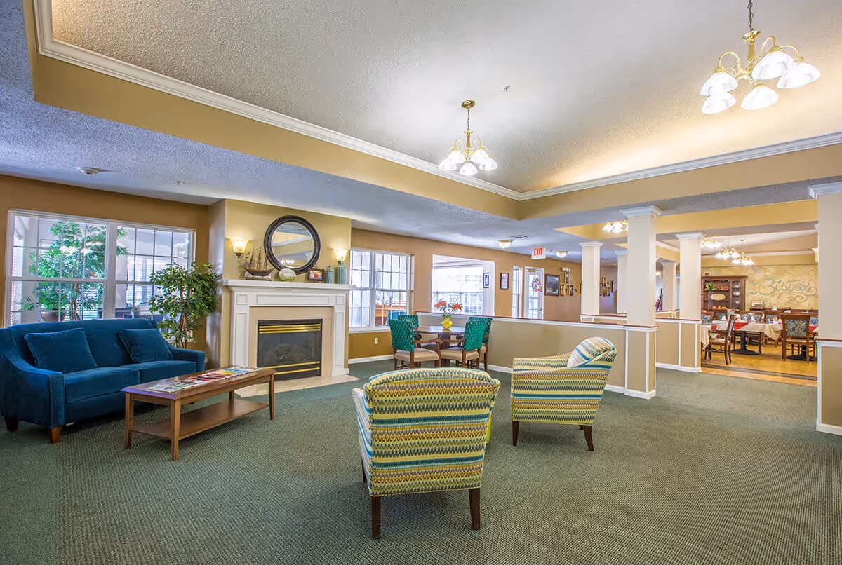 A spacious and well-lit common area in a senior living facility featuring a blue sofa, two patterned armchairs, a wooden coffee table, and a fireplace with a round mirror and decorative items on the mantel. There are large windows letting in natural light, a small table with four green chairs, and a dining area visible in the background with tables and chairs set up.