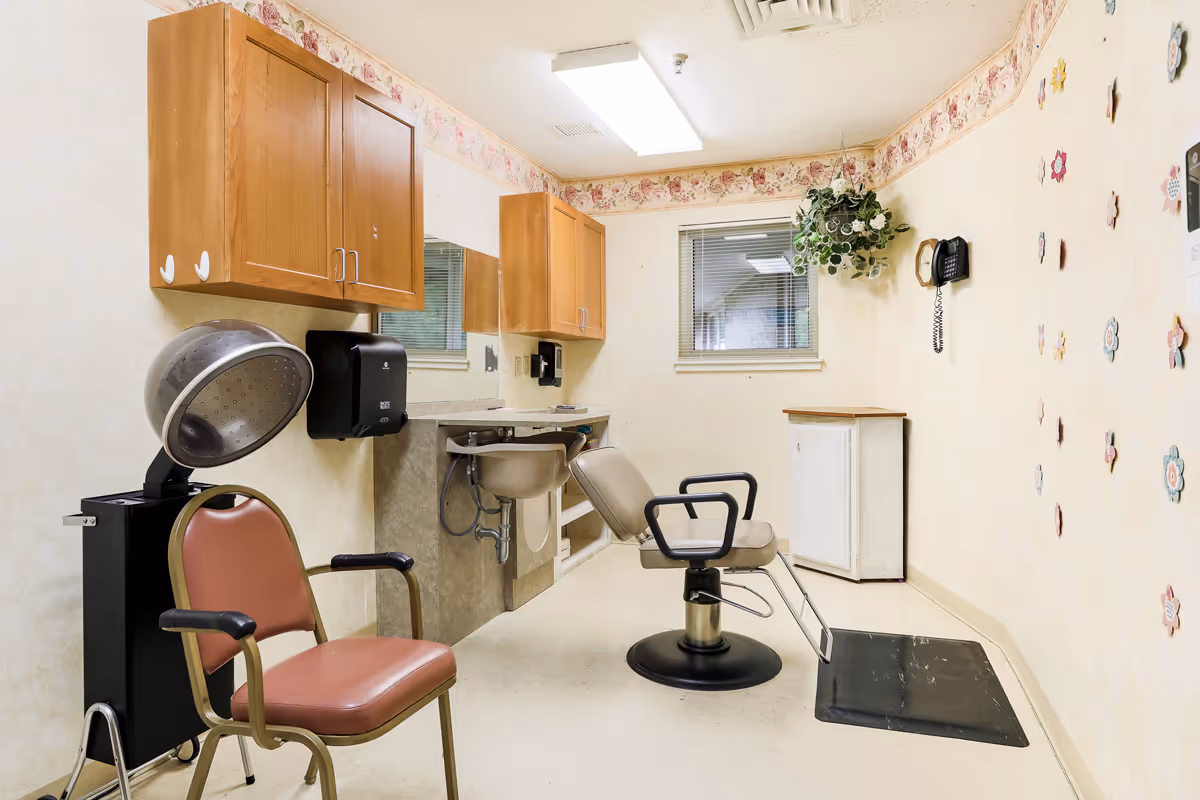 Small salon room with a hairdressing chair, hooded dryer, shampoo sink, cabinets and floral border wallpaper.
