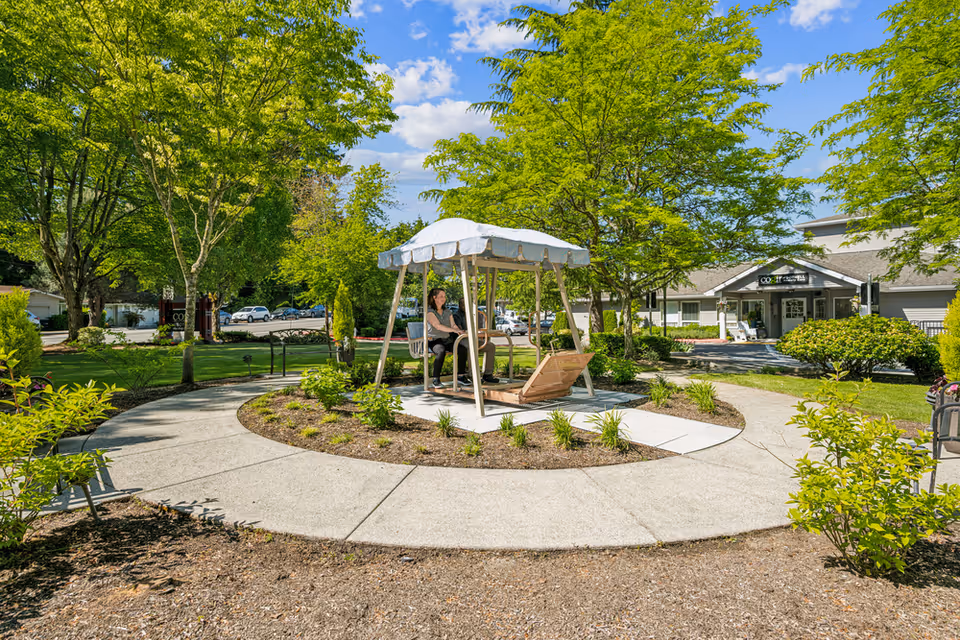 A sunny outdoor garden area at Cogir of Bothell Memory Care featuring a circular concrete pathway surrounding a small garden bed with plants. In the center, there is a wooden swing bench with a white canopy, where two people are seated. The background shows green trees, parked cars, and the entrance to the facility building.