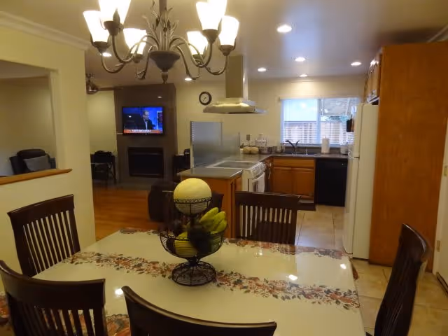 View of a dining area with a table covered by a floral tablecloth and a fruit basket centerpiece. The dining area opens into a kitchen with wooden cabinets, a white refrigerator, a stove, and a dishwasher. In the background, there is a living room with a wall-mounted TV above a fireplace and a chair visible.