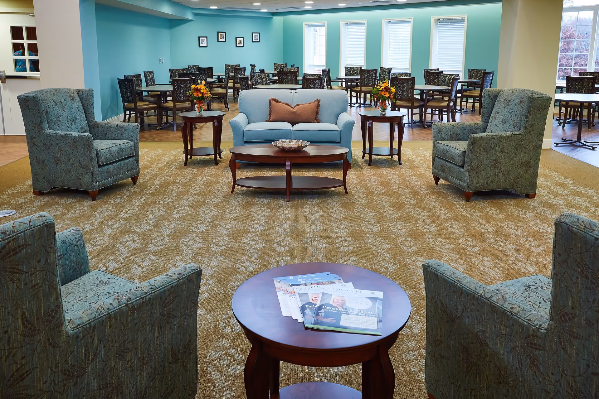 A spacious common area in a senior living facility with a seating arrangement consisting of a light blue loveseat with a brown pillow, four patterned armchairs, and three wooden coffee tables. The tables have flower vases and magazines on them. In the background, there are multiple dining tables and chairs against a teal-colored wall with framed pictures and windows letting in natural light.