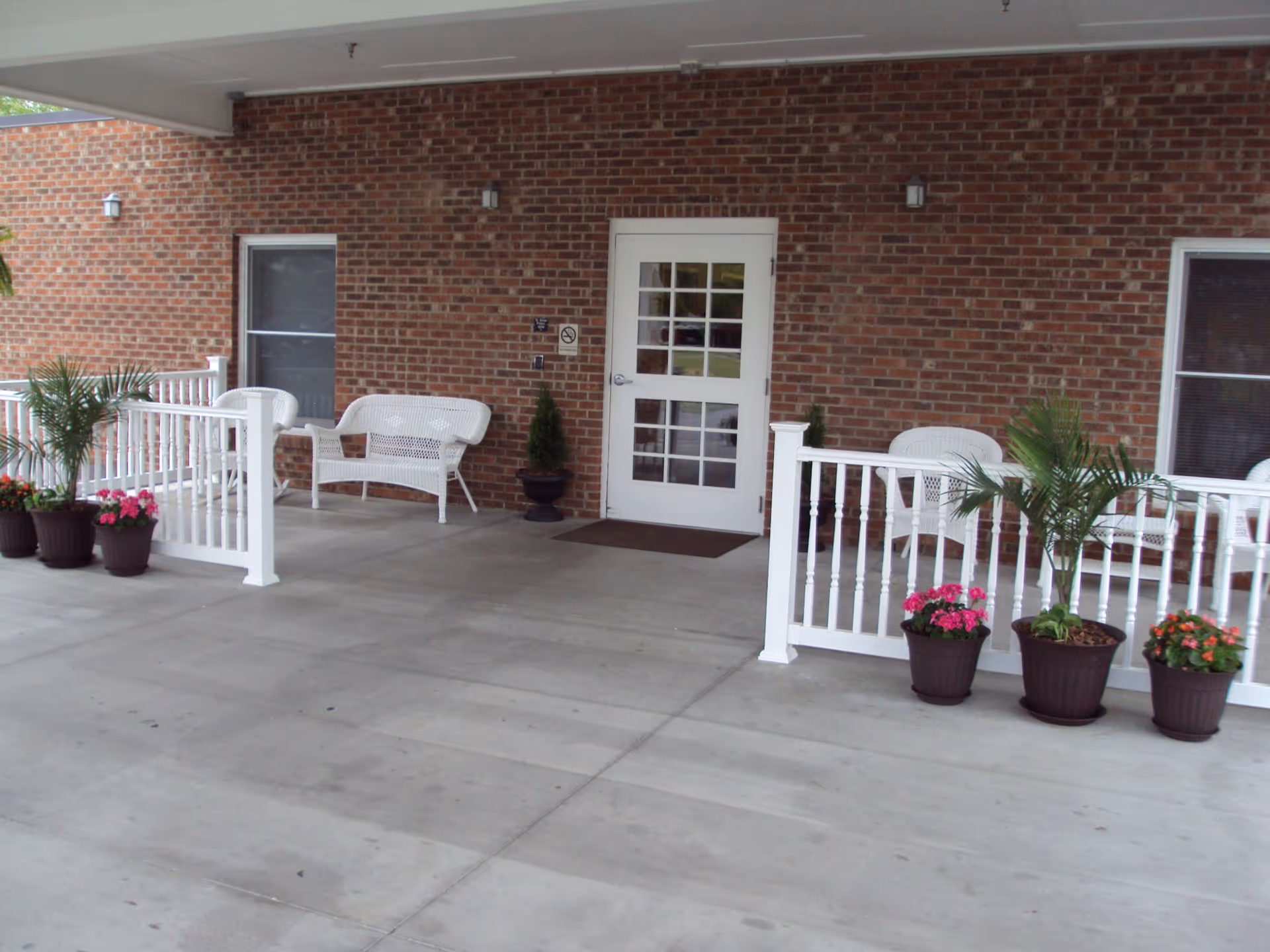 Covered outdoor patio area with white railings and white wicker chairs and benches. Several potted plants with green foliage and pink flowers are placed along the railing. The background features a brick wall with a white door and two windows.