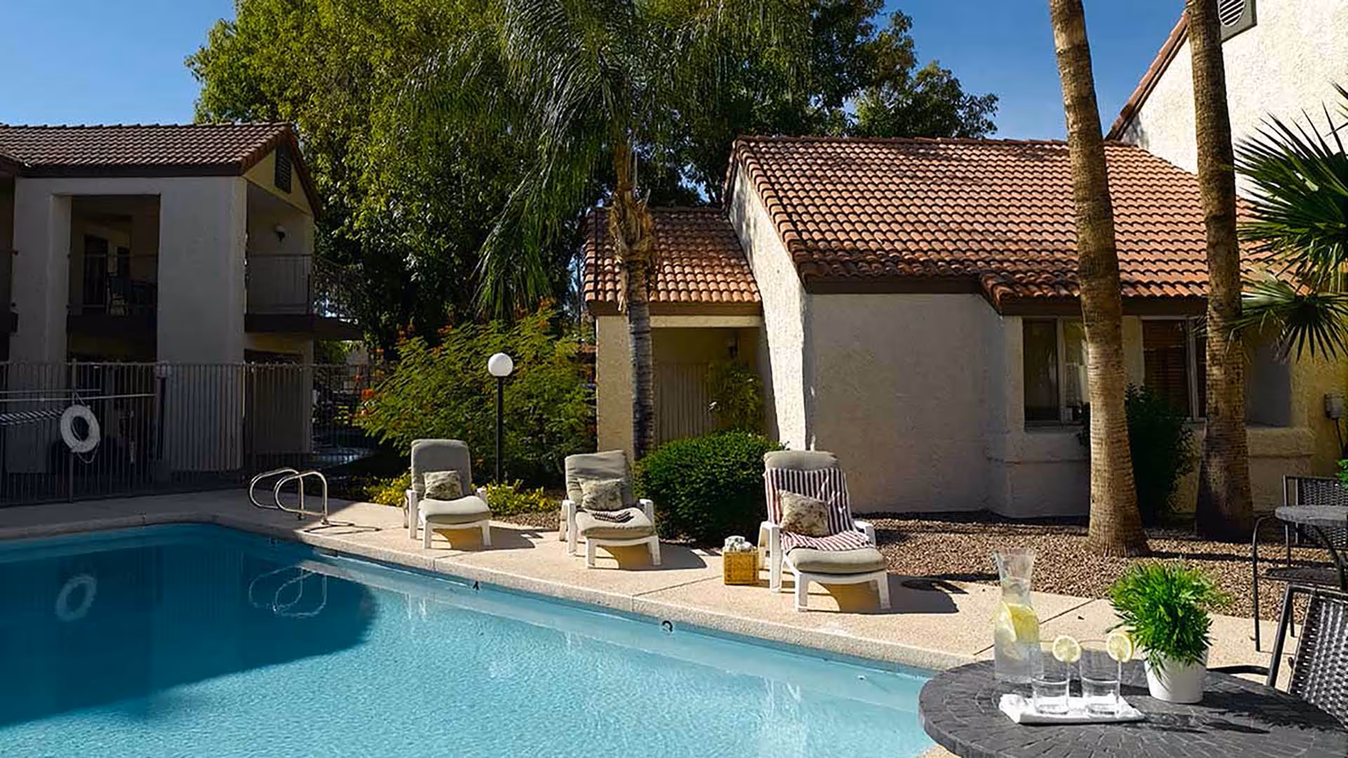 Outdoor pool area with clear blue water, surrounded by a concrete deck with four lounge chairs, some with cushions and towels. There are palm trees and bushes around, with beige stucco buildings with red tile roofs in the background. A round table with a pitcher of water, glasses, and a small potted plant is in the foreground.