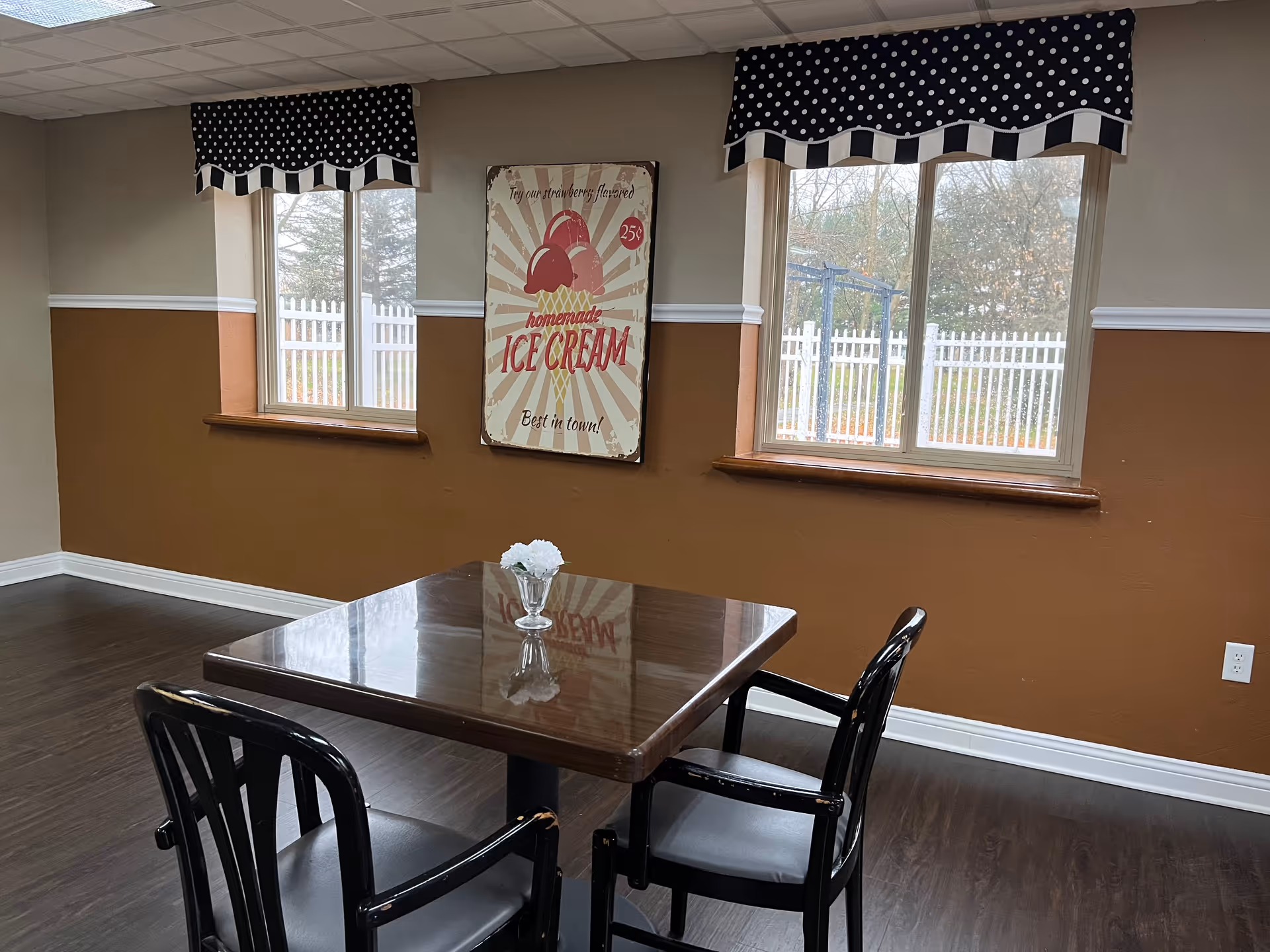 A small dining area with a square wooden table and two black chairs. On the table is a small glass vase with white flowers. The room has two windows with black and white polka dot valances, and a vintage-style wall sign advertising homemade ice cream for 25 cents. Outside the windows, a white picket fence and trees are visible.