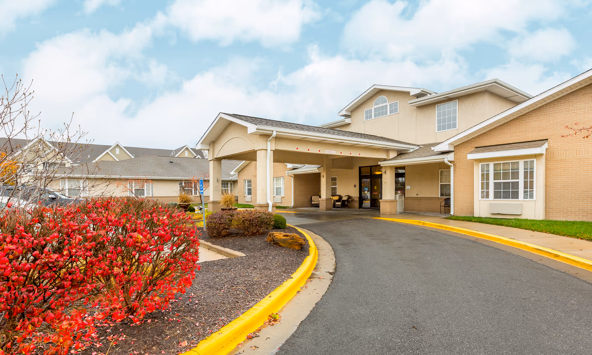 Exterior view of Brookdale Rosehill senior living facility showing the main entrance with a covered drop-off area, beige brick and stucco walls, and a curved driveway. There are red bushes and landscaping along the driveway, and a partly cloudy sky in the background.