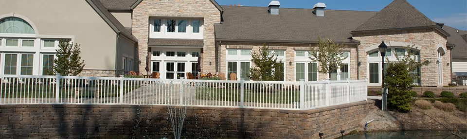 Exterior view of a senior living facility building with stone and beige walls, multiple windows, a white fence enclosing a small patio area with chairs, and a water fountain in a pond in the foreground.