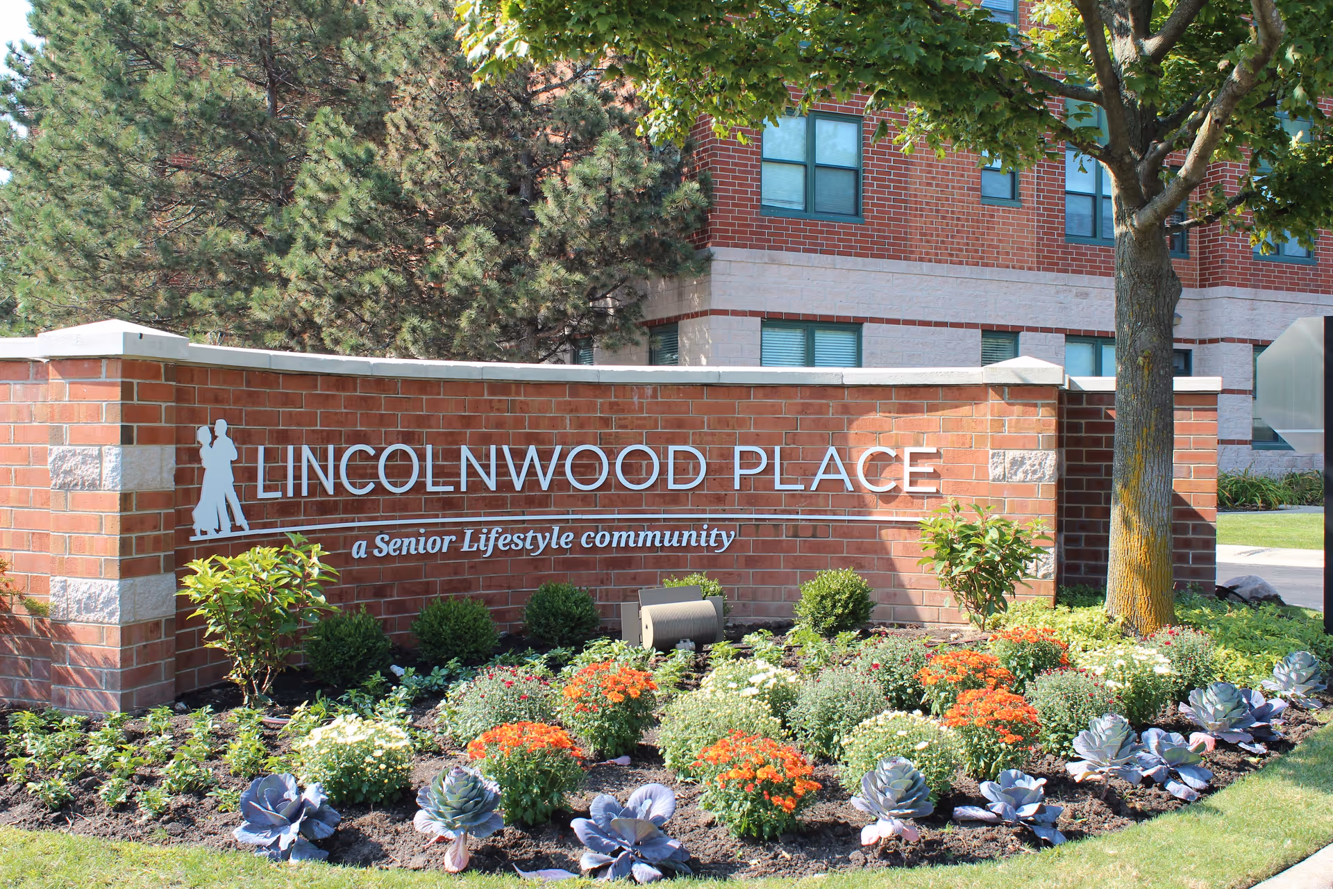 Brick sign for Lincolnwood Place, a senior lifestyle community, surrounded by a landscaped garden with various plants and flowers, with part of a brick building and trees in the background.