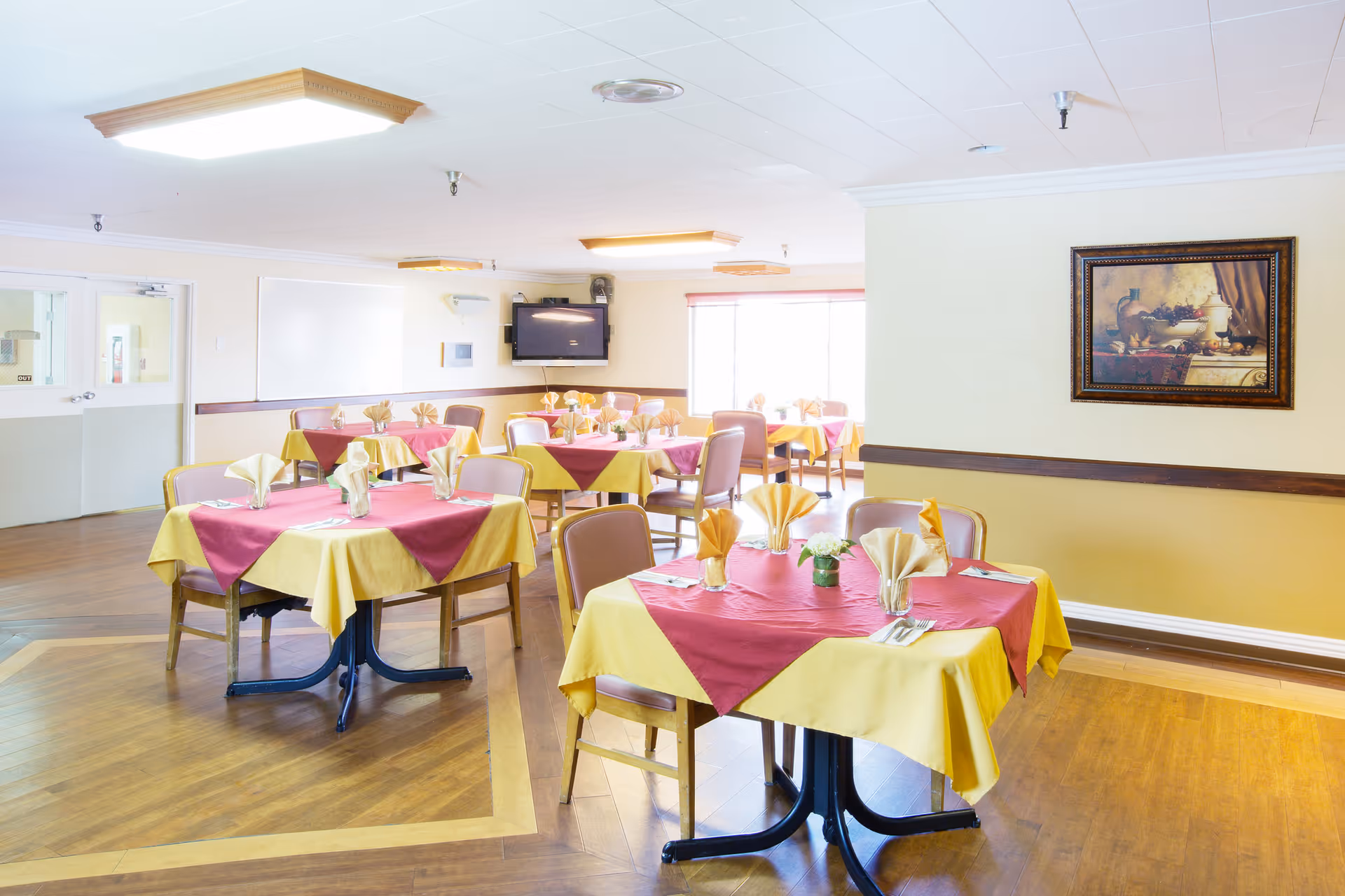 A bright dining room with multiple tables covered in yellow tablecloths with maroon overlays. Each table is set with folded napkins, utensils, and small flower arrangements. The room has wooden flooring, beige walls, a wall-mounted TV, and a framed painting on the wall. Large windows allow natural light to fill the space.