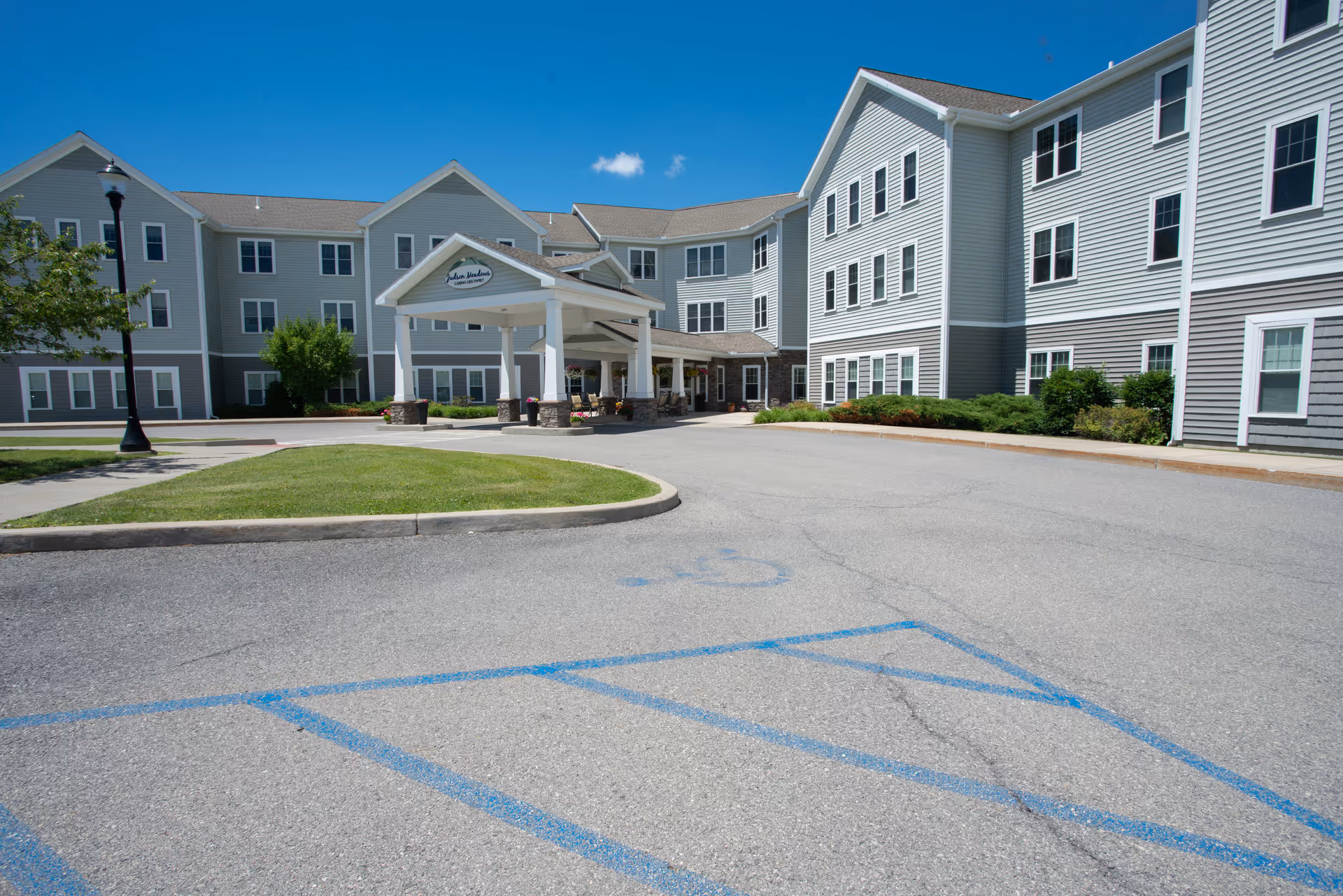 Front entrance and porte-cochère of a three-story gray assisted living building with a driveway and clear blue sky.