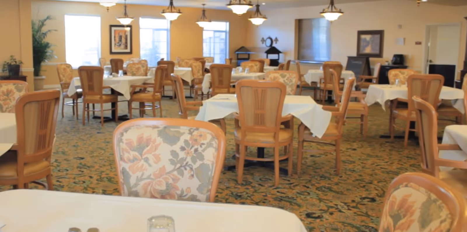 A spacious dining room with multiple round tables covered with white tablecloths. Each table is surrounded by wooden chairs with floral upholstered backs and seats. The room has a patterned carpet, several hanging light fixtures, framed artwork on the walls, and windows letting in natural light.
