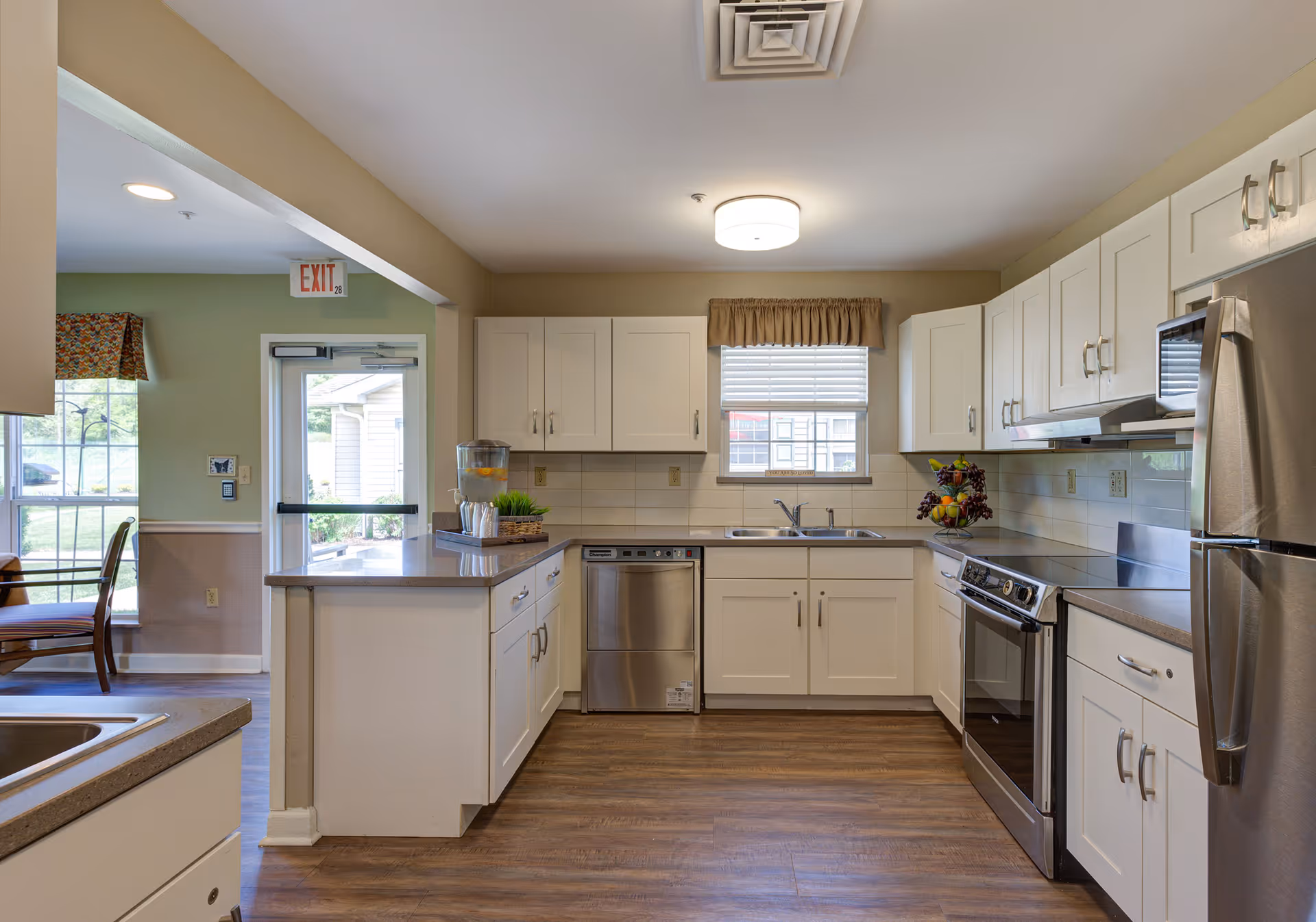 A clean and modern kitchen in a senior living facility with white cabinets, stainless steel appliances including a refrigerator, dishwasher, and stove. There is a window above the sink with a beige valance, a countertop with a water dispenser and a small plant, and wood-look flooring. To the left, there is a partial view of a dining area with chairs and a window.