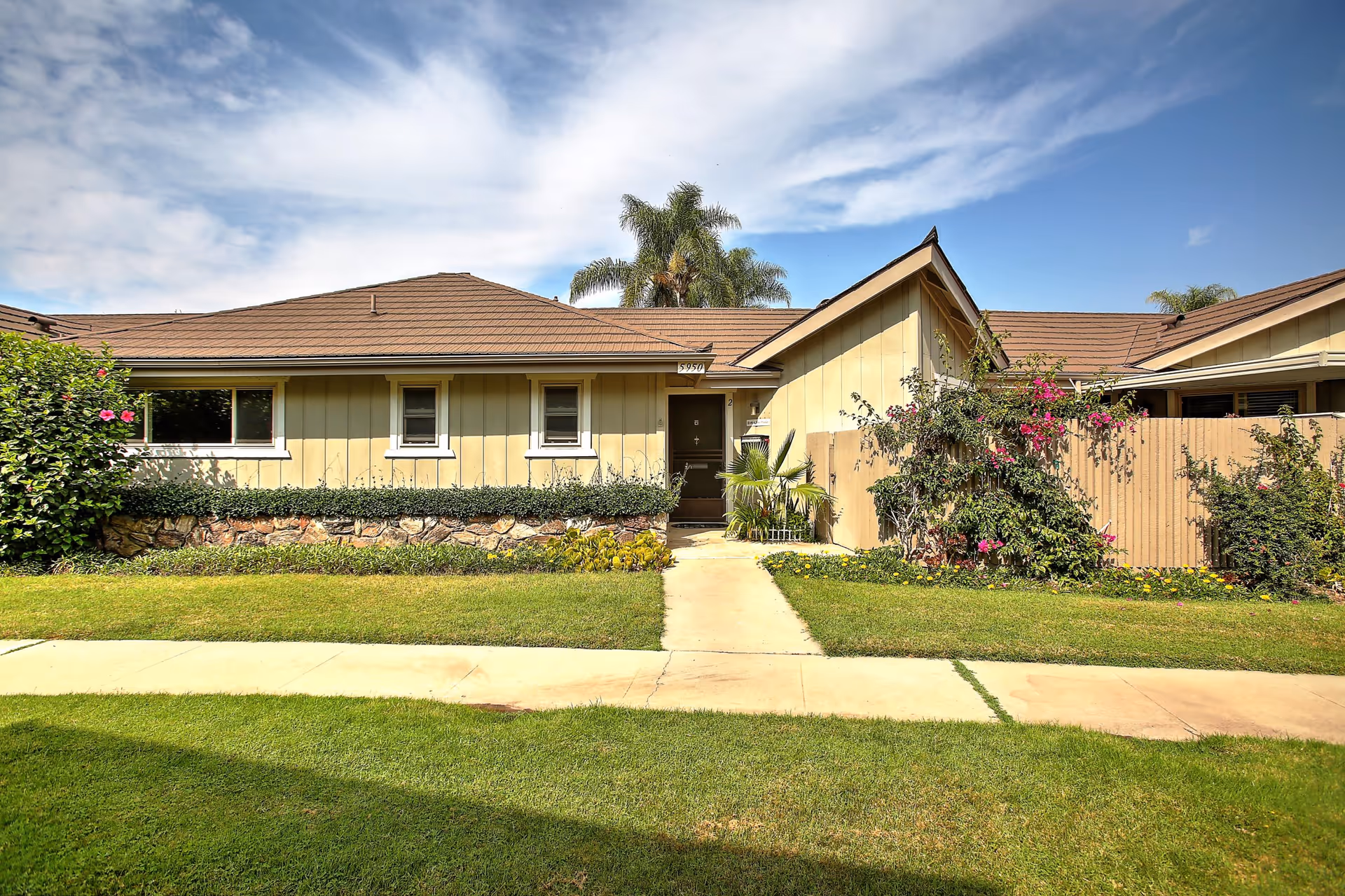 Exterior view of a single-story residential building with a brown shingled roof, beige siding, and a small front garden with green grass, bushes, and flowering plants. A concrete walkway leads to a dark brown front door under a small porch. Palm trees are visible in the background under a partly cloudy blue sky.