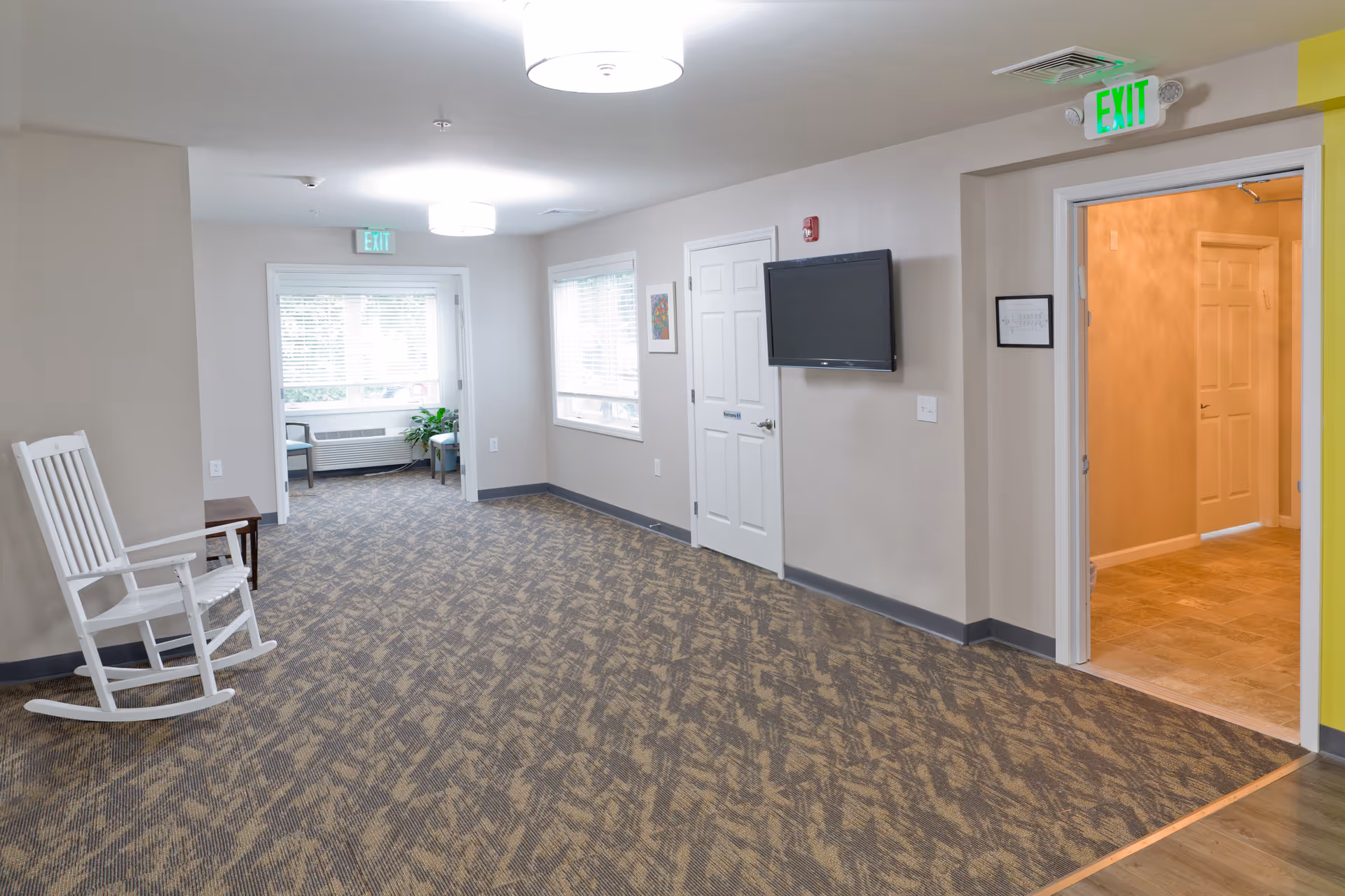 Carpeted common area in an assisted living facility with a white rocking chair, wall-mounted TV, seating by windows, and exit signs.