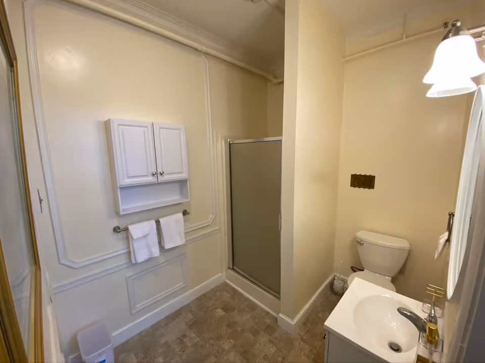 A clean bathroom with beige walls and tiled floor. The room features a white toilet, a white sink with a faucet, a mirror with a light fixture above it, a small shower with a frosted glass door, a wall-mounted cabinet with two doors, and two white towels hanging on a towel rack beneath the cabinet.