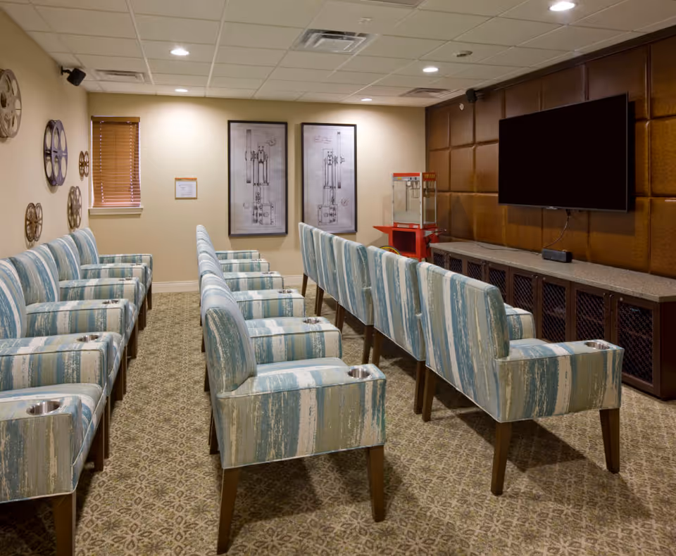A small theater room with two rows of blue and white striped upholstered chairs facing a large flat-screen TV mounted on a brown padded wall. There is a popcorn machine on a red stand in the corner, patterned carpet flooring, and framed artwork on the far wall.
