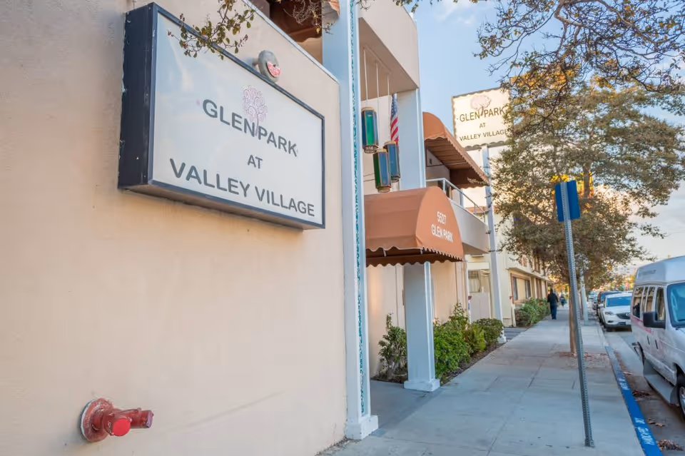 Exterior view of Glen Park at Valley Village facility showing the building entrance with an awning, a sign with the facility name, a sidewalk with trees, parked cars, and a person walking in the distance.