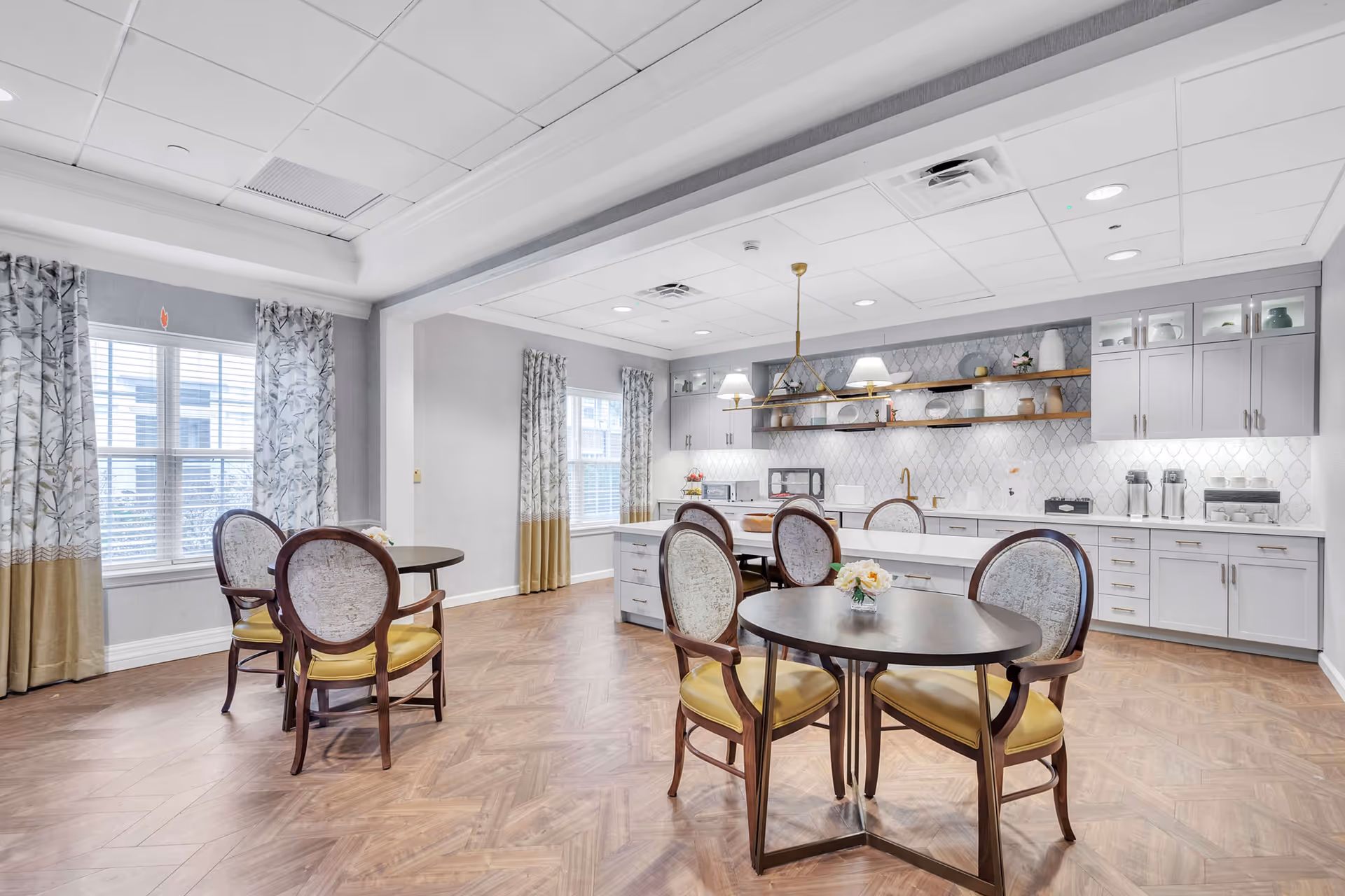 Bright communal dining room with round tables and upholstered chairs facing a modern kitchenette with white cabinets and open shelving.