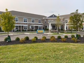 Front exterior view of a two-story senior living facility building with a covered entrance, multiple windows, and landscaped greenery including trees and shrubs in front.
