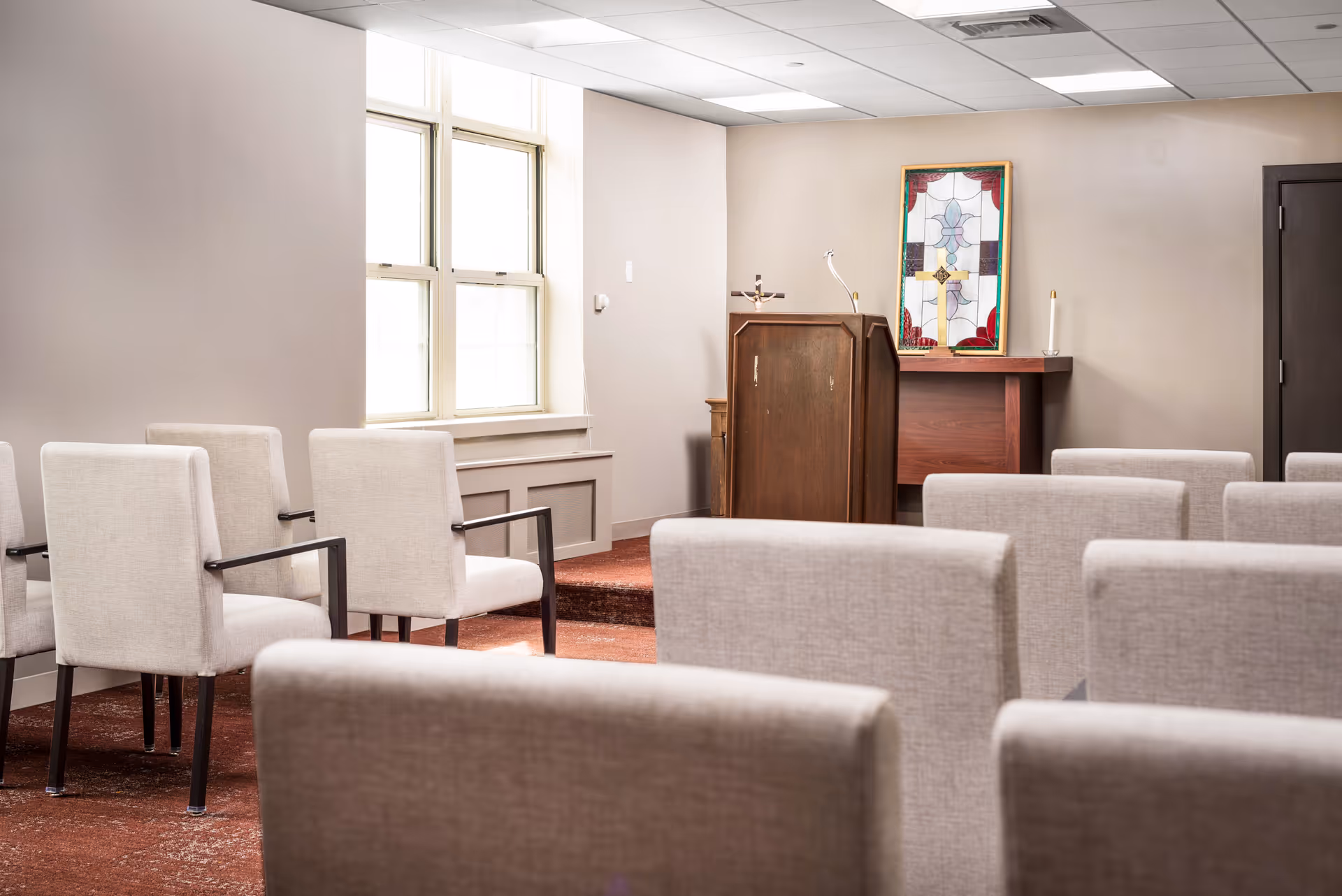 Interior view of a small chapel or meditation room with rows of beige upholstered chairs facing a wooden podium and a table with a stained glass artwork on the wall behind it.