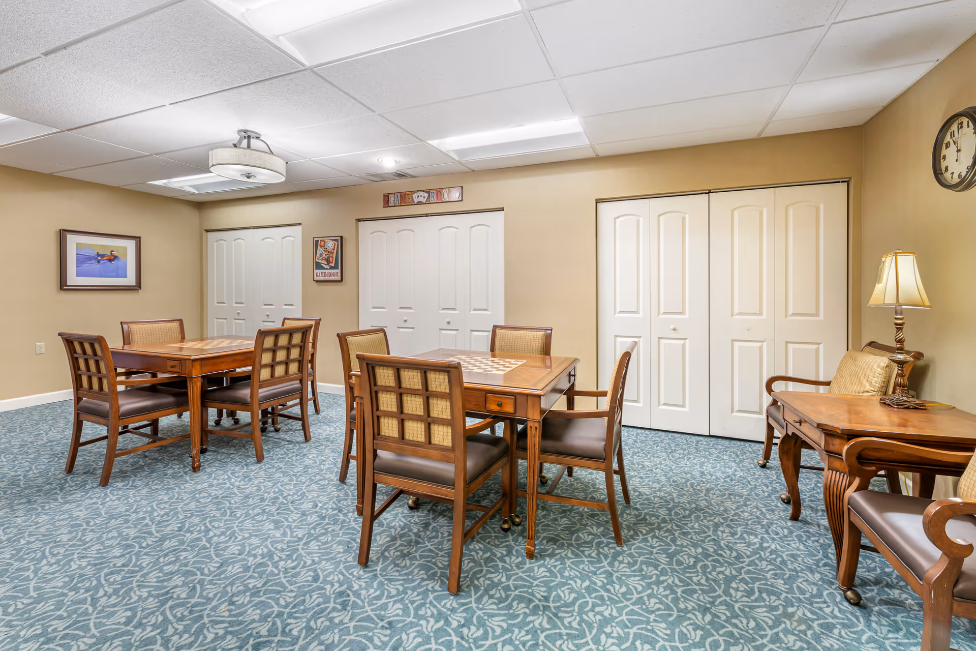 Carpeted communal room with several wooden tables and chairs, side tables and lamps against beige walls and closed closet doors.