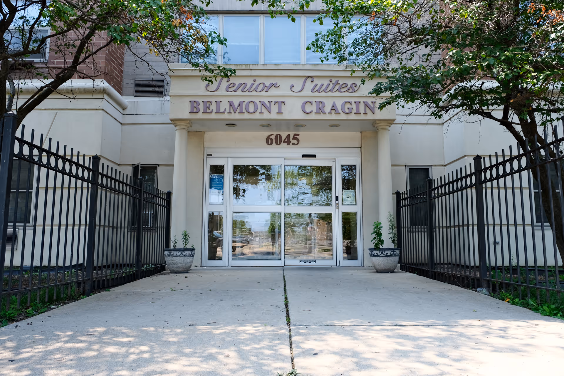 Entrance to Senior Suites Belmont Cragin building with glass double doors, flanked by two potted plants and black metal fencing on both sides. The building number 6045 is displayed above the doors under the facility name.