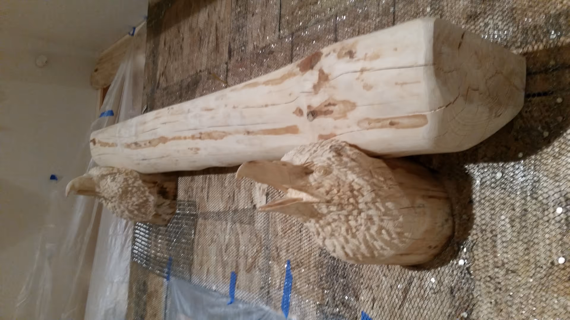 Two wooden sculptures of birds placed on a textured surface indoors, with a large wooden beam behind them and plastic sheeting taped to the wall in the background.